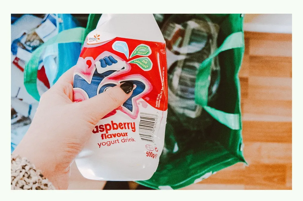 Mano che tiene una bottiglia di yogurt di plastica vuota sopra una borsa verde per il riciclaggio Mano che tiene una bottiglia di yogurt di plastica vuota sopra una borsa verde per il riciclaggio